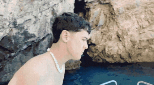 A Shirtless Man Wearing A Pearl Necklace Is Standing In Front Of A Body Of Water GIF