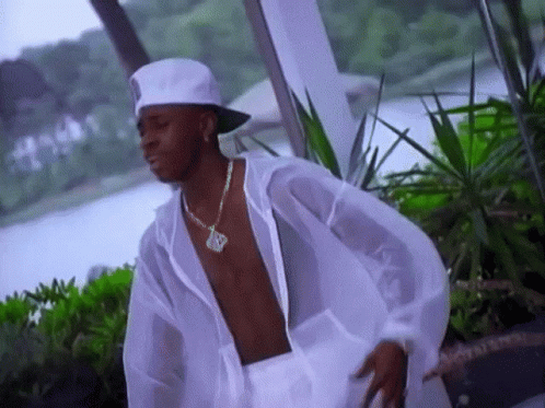 A Shirtless Man Wearing A White Hat And A Necklace With The Letter R On It Is Standing In Front Of A Body Of Water GIF