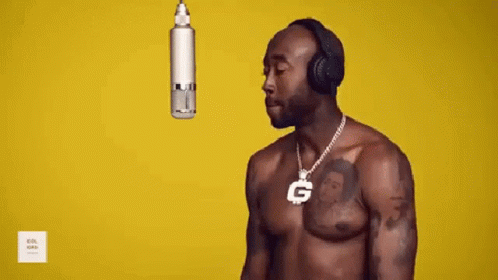 A Shirtless Man Wearing Headphones And A Necklace With A G On It Is Standing In Front Of A Microphone . GIF