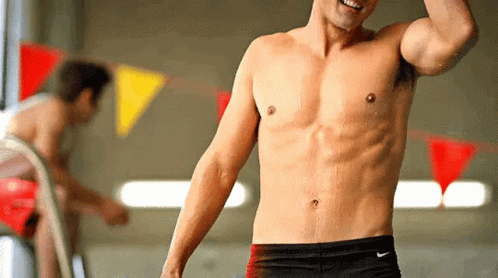 A Shirtless Man Wearing Nike Swim Trunks Stands In Front Of A Swimming Pool GIF