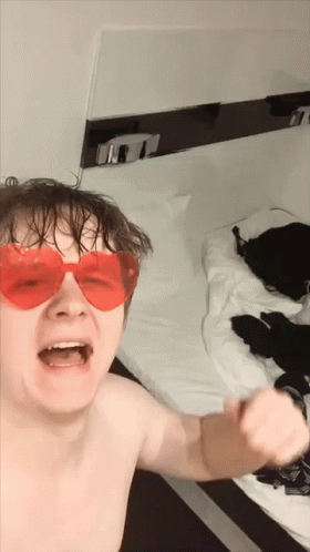 A Shirtless Man Wearing Red Heart Shaped Sunglasses Is Standing In Front Of A Bed GIF