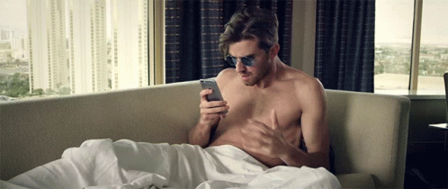 A Shirtless Man Wearing Sunglasses Is Laying On A Couch Looking At His Phone GIF