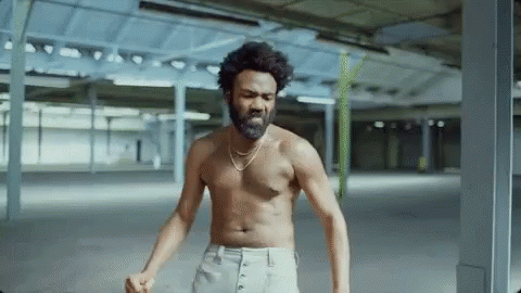 A Shirtless Man With A Beard And A Necklace Is Standing In A Warehouse . GIF