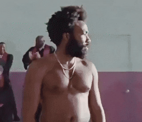 A Shirtless Man With A Beard And A Necklace Is Standing In Front Of A Group Of Men . GIF