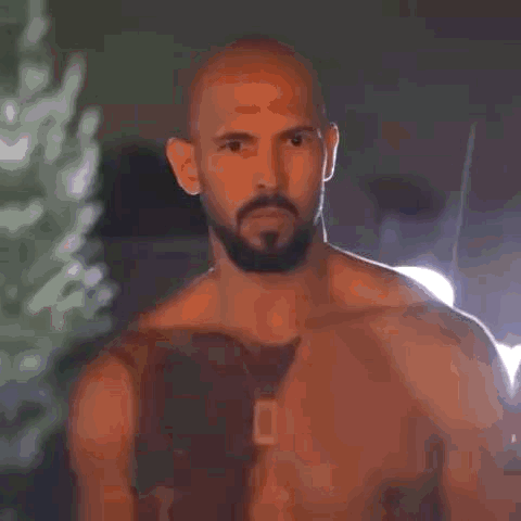 A Shirtless Man With A Beard And A Tattoo On His Chest Is Standing In Front Of A Building . GIF