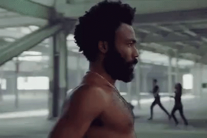 A Shirtless Man With A Beard And Afro Is Standing In A Building . GIF