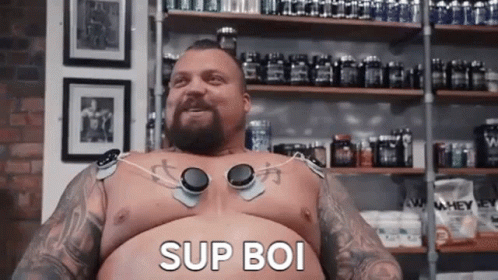 A Shirtless Man With A Beard And Tattoos Is Sitting In Front Of A Shelf With Bottles Of Supplements . GIF