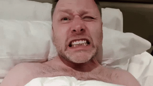 A Shirtless Man With A Beard Is Making A Funny Face While Laying In Bed GIF
