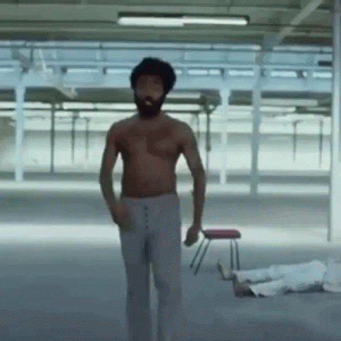 A Shirtless Man With A Beard Is Standing In A Room With A Chair In The Background . GIF