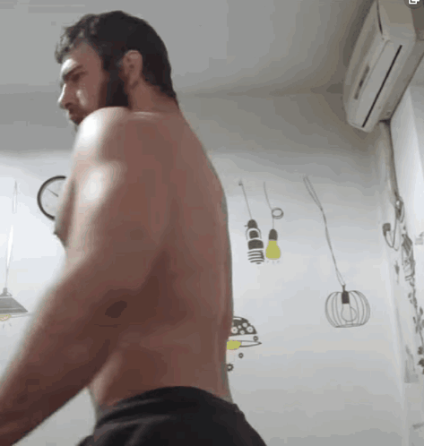 A Shirtless Man With A Beard Is Standing In A Room With A Clock On The Wall . GIF
