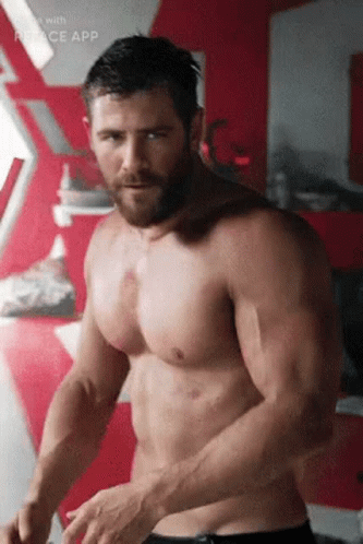 A Shirtless Man With A Beard Is Standing In Front Of A Red Wall In A Room . GIF