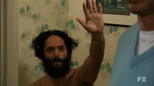 A Shirtless Man With A Beard Is Waving His Hand In Front Of A Wall . GIF