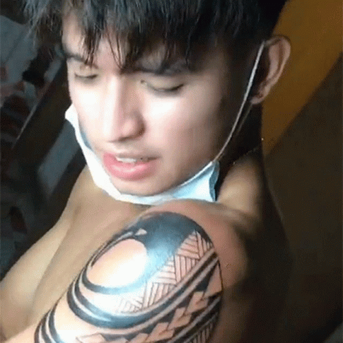 A Shirtless Man With A Mask On His Face Is Getting A Tattoo On His Arm . GIF