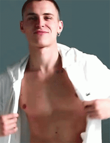 A Shirtless Man With A Tattoo On His Chest Is Taking Off His Shirt And Smiling . GIF