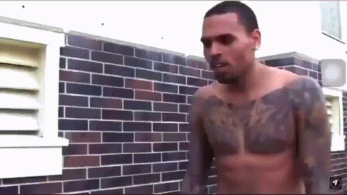 A Shirtless Man With A Tattoo On His Chest Stands In Front Of A Brick Wall GIF