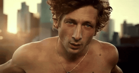 A Shirtless Man With Curly Hair Wearing A Necklace Looks At The Camera GIF
