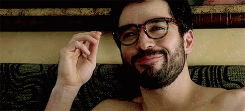 A Shirtless Man With Glasses And A Beard Is Laying On A Bed . GIF