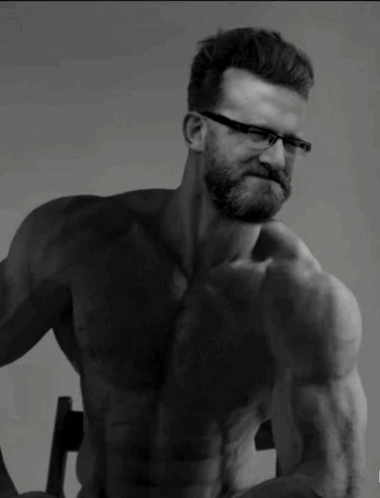 A Shirtless Man With Glasses And A Beard Is Sitting In A Chair GIF