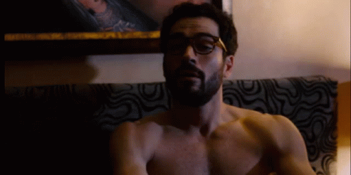 A Shirtless Man With Glasses And A Beard Is Sitting On A Couch GIF