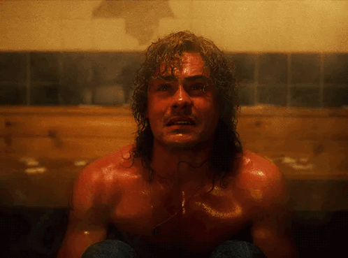A Shirtless Man With Long Hair Is Sitting In A Bathroom GIF