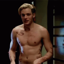 A Shirtless Man With Tattoos On His Arm And Chest Is Standing In Front Of A Door . GIF