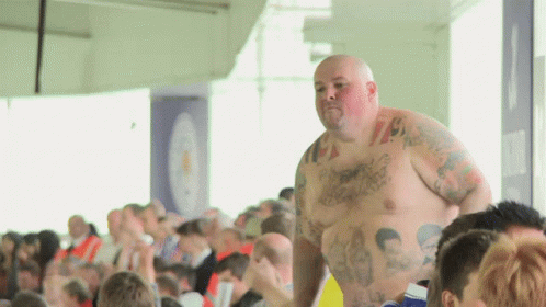 A Shirtless Man With Tattoos On His Chest Stands In Front Of A Crowd Of People GIF