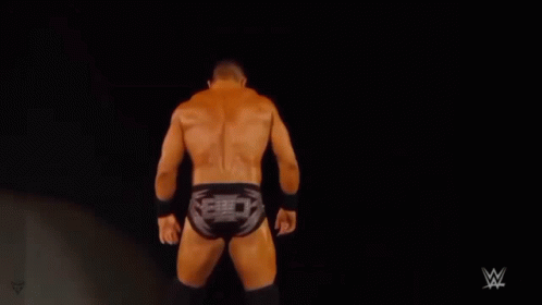 A Shirtless Wrestler Is Standing On A Stage In Front Of A Black Background . GIF