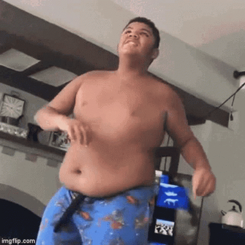 A Shirtless Young Man Is Dancing In A Living Room While Wearing Blue Shorts . GIF