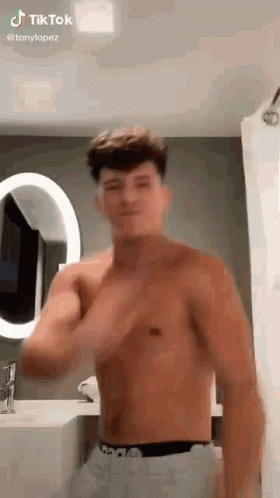 A Shirtless Young Man Is Standing In Front Of A Mirror In A Bathroom . GIF