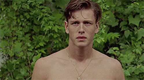 A Shirtless Young Man Wearing A Gold Necklace Is Standing In Front Of A Bush . GIF