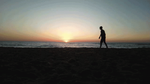 A Silhouette Of A Man Walking On A Beach At Sunset GIF