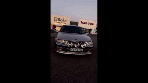 A Silver Car Is Parked In Front Of A Building That Says Toys Too GIF