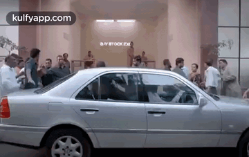 A Silver Car Is Parked In Front Of A Building With A Crowd Of People Standing Around It . GIF