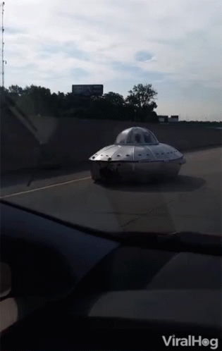 A Silver Flying Saucer Is Driving Down A Highway With A Billboard In The Background GIF