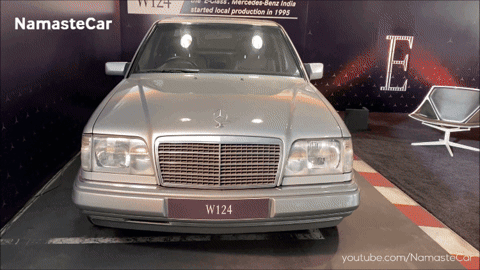 A Silver Mercedes W124 Is On Display In Front Of A Sign That Says Namaste Car GIF