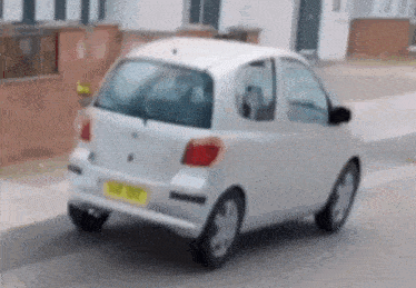 A Small White Car Is Parked On The Side Of The Road With A Yellow Plate GIF