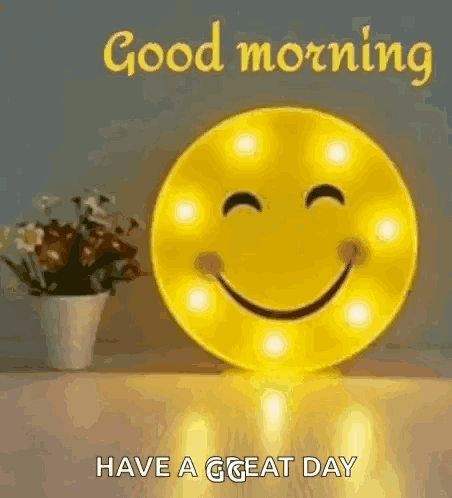 A Smiley Face Is Sitting On A Table Next To A Vase Of Flowers And Says `` Good Morning Have A Great Day '' . GIF