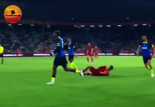 A Soccer Player In A Blue Jersey Is Running Towards A Player In A Yellow Jersey Who Is Laying On The Ground GIF
