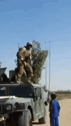 A Soldier Is Standing On Top Of A Military Vehicle While A Boy Looks On . GIF