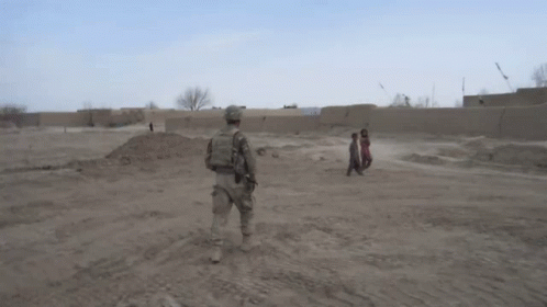a soldier stands in the middle of a dirt field with two children walking behind him