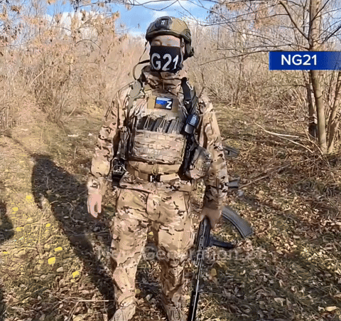A Soldier Wearing A Mask With G21 On It Stands In The Woods GIF