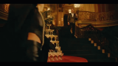 A Stack Of Champagne Glasses On A Table With A Woman Standing In The Background GIF