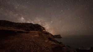 A Starry Night Sky Over A Body Of Water With A Cliff In The Foreground GIF