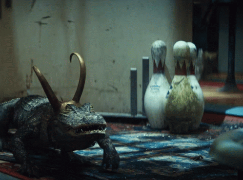 A Statue Of A Crocodile With A Crown On Its Head Stands Next To Bowling Pins GIF