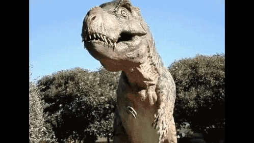 A Statue Of A Dinosaur Is Standing In Front Of Some Trees GIF
