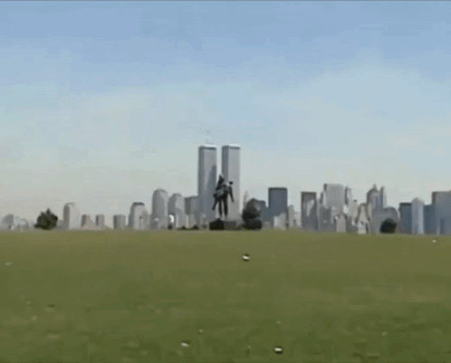 A Statue Of A Man Carrying Another Man In Front Of The Twin Towers Of The World Trade Center GIF