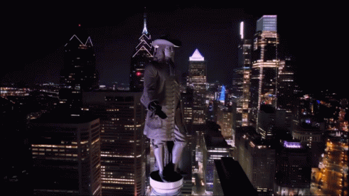 A Statue Of A Man In A Hat Stands On Top Of A City Skyline At Night GIF
