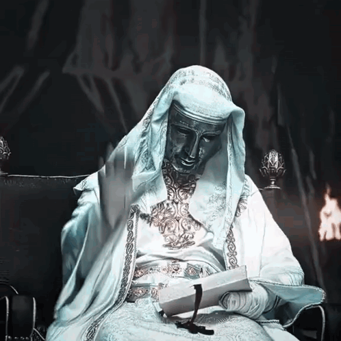 A Statue Of A Man In A White Robe Sits In A Chair Reading A Book GIF