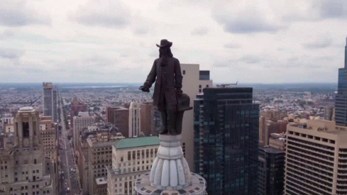 A Statue Of A Man Stands On Top Of A Building In A City With A Chase Bank Building In The Background GIF