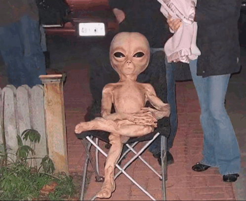 A Statue Of An Alien Is Sitting In A Chair With A License Plate That Says A1 GIF
